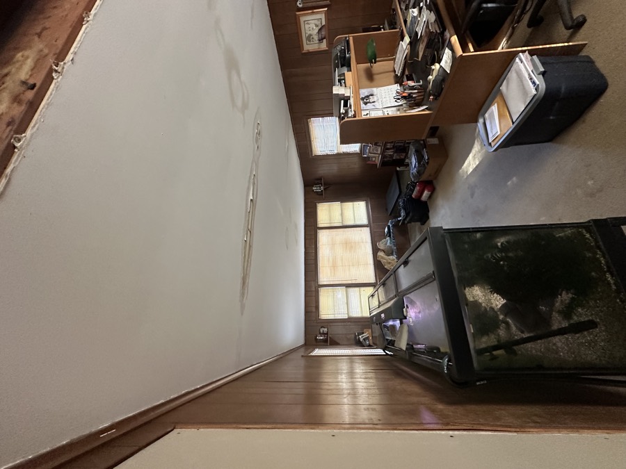 A wood-panelled room with a water-stained ceiling, an exposed joist where the ceiling has been opened up, and a lifetime of furniture and belongings still in place.