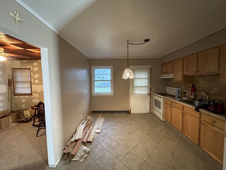 A dated kitchen and dining room with original cabinets and vinyl flooring, mid-clear-out, with flooring materials stacked on the floor and packing boxes in the next room.